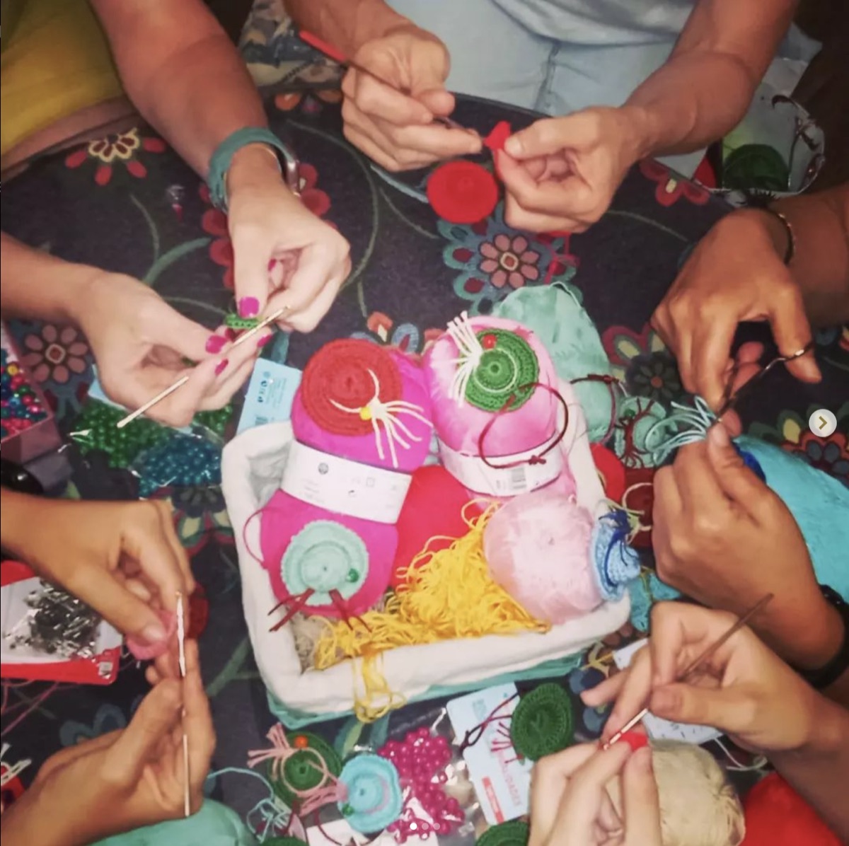 Varias manos tejiendo pequeños sombreros de ganchillo de colores alrededor de una cesta de lana